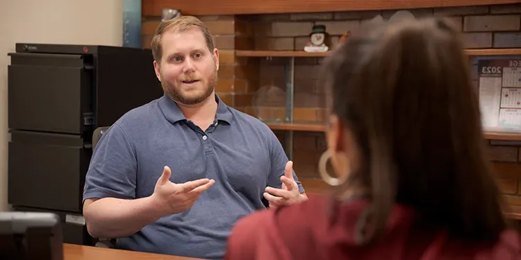 Two people discussing something. The image is a closeup of the back of a woman's shoulder while she talks to a man